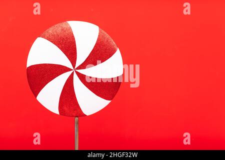 Rot-weißer Lollipop wirbelt auf einem weißen Stock über rotem Hintergrund. Draufsicht, Kopierraum. Riesige rote und weiße Süßigkeiten. Süße Stockfoto