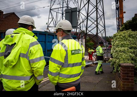 Arbeiter in der PSA verwenden oxyde Acetylenbrenner, um beschädigte Teile eines geerdeten Strommasylons auszuschneiden, während Techniker zuschauen Stockfoto