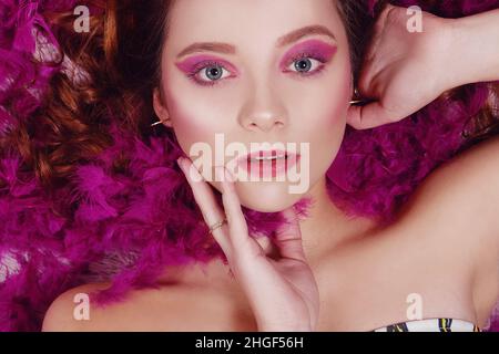 Ein unglaublich schönes Mädchen mit rosa Mode Make-up liegt in Federn. Ein Mädchen mit perfekten Augenbrauen und Make-up schaut auf die Kamera. Helle Augen Stockfoto