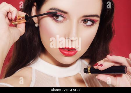 Portrait, Model hält Mascara in den Händen und malt Wimpern. Schöne Brünette mit professionellem roten Make-up, auf rotem Hintergrund. Die rote Neujahrsgnade Stockfoto