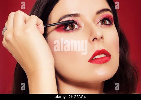 Portrait, Model hält Mascara in den Händen und malt Wimpern. Schöne Brünette mit professionellem roten Make-up, auf rotem Hintergrund. Die rote Neujahrsgnade Stockfoto