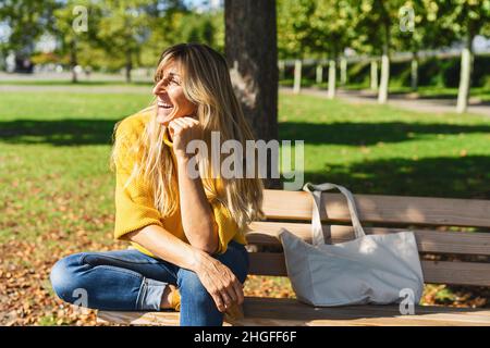 Glückliche blonde Frau im Freien sitzen und denken. Frau entspannen auf einer Holzbank mit Leinwand Stofftasche unter dem Baum in einem Sommertag. Porträt von w Stockfoto