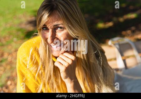 Porträt einer schönen Frau, die in einem Stadtpark sitzt. Fröhliche lächelnde Frau, die im Sommer auf einer Bank im Freien sitzt und die Kamera anschaut. Porträt der sorglosen w Stockfoto