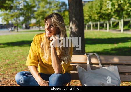 Lächelnde blonde Frau sitzt im Freien und denkt. Frau entspannt auf einer Holzbank mit Stoff Stoff Tasche unter dem Baum in einem Sommertag. Porträt von Stockfoto