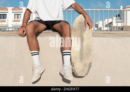 Nicht erkennbarer Skater, der am Rand der Skaterampe sitzt. Er trägt Sommerkleidung. Stockfoto