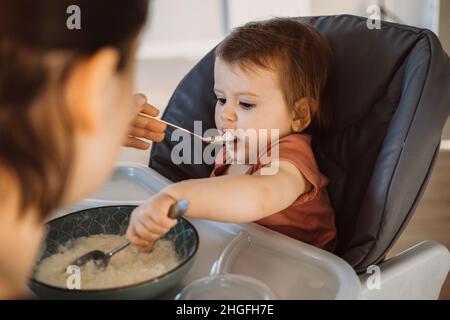Baby, das auf einem Hochstuhl sitzt und von den Händen der Mutter gefüttert wird. Für Lifestyle-Design. Gesunde Ernährung. Stockfoto