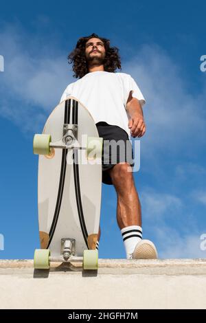 Junger, hübscher Skater an der Spitze der Skaterampe. Low-Angle-Aufnahme. Er hat lange schwarze Haare und einen Bart. Er trägt Sommerkleidung. Stockfoto