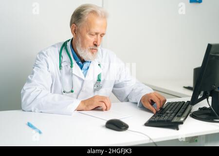 Porträt eines fokussierten reiferen männlichen Arztes mit weißem Mantel und Stethoskop, der am Desktop-Computer am Schreibtisch im medizinischen Büro sitzt. Stockfoto