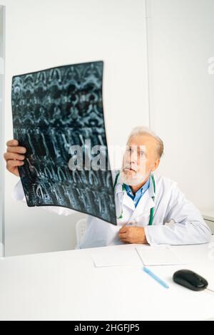 Vertikale Aufnahme eines erwachsenen Arztes, der die MRT-Wirbelsäulenaufnahme eines Patienten untersucht, der am Schreibtisch im medizinischen Bürozimmer sitzt. Stockfoto