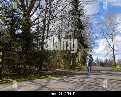 Woodinville, WA USA - ca. April 2021: Blick auf einen älteren Mann in einem Baseballhut und Wasserläufer, der an einem hellen, sonnigen Tag auf einem gepflasterten Radweg entlang geht. Stockfoto