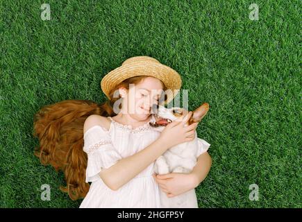 Nettes Mädchen und Hund umarmen, küssen und liegen auf dem grünen Gras im Frühlingspark. Frühling, Ostern, Freunde Konzept. Stockfoto