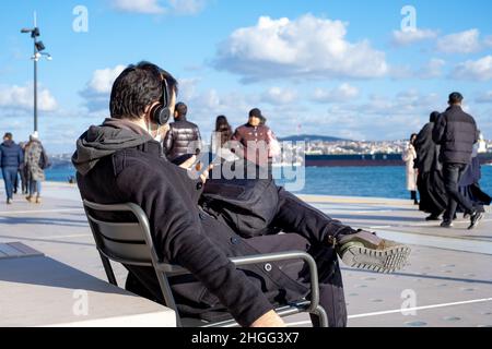 Mann, der am Meer sitzt und auf sein Telefon schaut. Mann, der am Meer sitzt und auf sein Telefon schaut, ist vor dem Hintergrund Meer und wolkenverhaufiger Himmel. Stockfoto