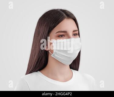 Modell einer weißen OP-Maske mit Ohrschlaufen auf einer dunkelhaarigen Krankenschwester, isoliert auf dem Hintergrund. Schützende Atemschutzmaske auf einem schönen kaukasischen Mädchen. Tem Stockfoto