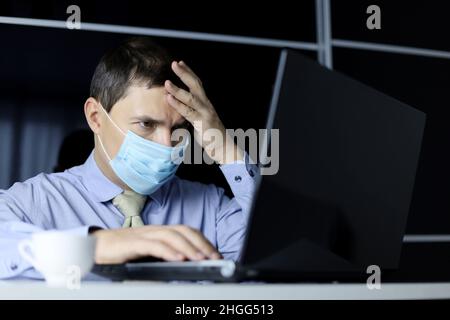 Mann in Gesichtsmaske und Bürokleidung, der auf das Notebook-Display fokussiert schaut. Konzept der Lösung einer schwierigen Aufgabe Stockfoto