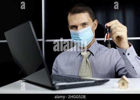 Immobilienmakler mit Wohnungsschlüsseln in den Händen. Mann in medizinischer Gesichtsmaske und Bürokleidung sitzt am Laptop, Konzept des Kaufs oder Vermietung zu Hause Stockfoto