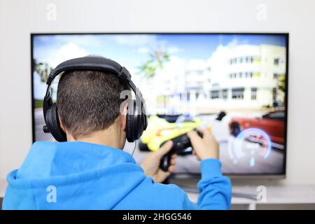 Kerl in Kopfhörern spielt mit einem Gamepad ein Rennvideospiel auf dem Fernsehbildschirm. Konzept der Spielsucht, Freizeit zu Hause Stockfoto