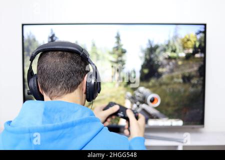 Kerl in Kopfhörern, der mit einem Gamepad-Shooter-Videospiel auf dem Fernsehbildschirm spielt. Konzept der Spielsucht, Freizeit zu Hause Stockfoto