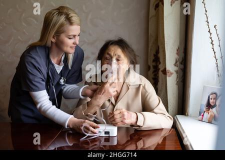 Junge hochqualifizierte angenehme Ärztin, die Verneblermaske aufsetzt Gesicht ihrer älteren weiblichen Patientin, um beim Einatmen zu machen Besucht ihn zu Hause Stockfoto