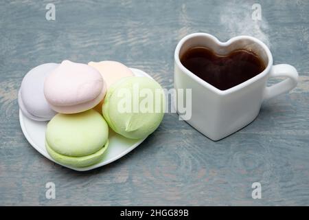 Runde mehrfarbige Marschwalben im Stapel und ein weißer herzförmiger Becher mit schwarzem dampfendem heißen Kaffee auf blauem Holzhintergrund. Frühstück für die Liebe Stockfoto