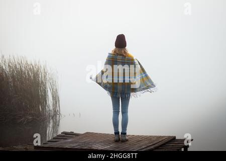 Frau in warme Decke gehüllt, die auf dem Pier am See steht. Weibliche Person in neblig kalten Herbstmorgen in der Natur entspannen. Camping im Freien Stockfoto