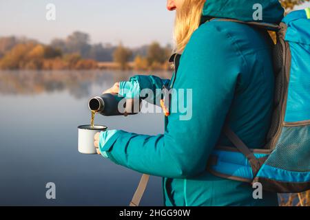 Die Frau gießt heißes Getränk aus Thermoskannen in den Reisebecher. Erfrischung beim Wandern am See im Herbst Stockfoto