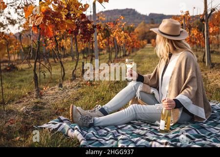 Frau mit Hut und Poncho trinkt Weißwein. Winzerin auf der Decke im Weinberg sitzend und in der Herbstsaison entspannt. Entspannung im Herbst Weingut Stockfoto