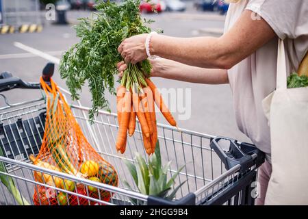 Einkaufen ohne Plastik. Frau, die auf dem Parkplatz Karotten aus dem Warenkorb holt. Wiederverwendbarer Netzbeutel. Zero Waste Konzept Stockfoto