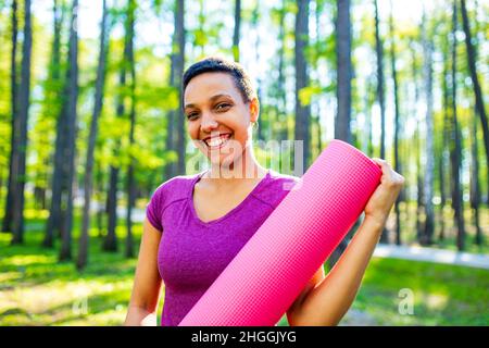Glückliche junge afroamerikanische Trainerin mit kurzen Haaren, die eine Yogamatte im Freien hält und die Kamera anschaut Stockfoto