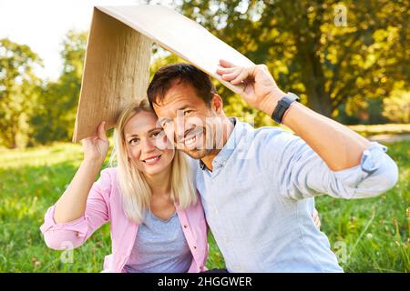 Glückliches junges Paar im Park mit Dach über dem Kopf, das den Besitz und die Sicherheit des Hauses symbolisiert Stockfoto
