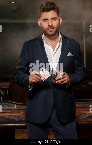 Selbstbewusster junger Mann mit zwei Assen und einem Glas Getränk in einem Glücksspielbetrieb Stockfoto