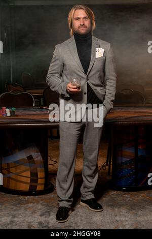 Selbstbewusster Mann mit zwei Assen in der Jackentasche und einem Glas Getränk in der Hand, der im Casino steht Stockfoto