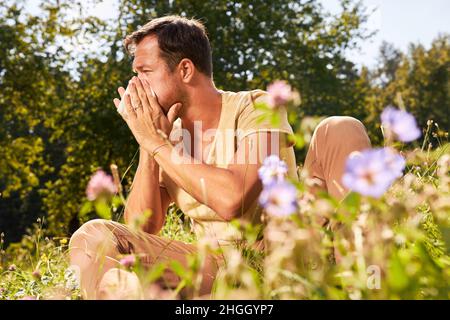 Mann mit Pollenallergie sitzt zwischen blühenden Blumen und bläst seine Nase Stockfoto