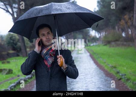 Junger eleganter Mann mit Regenschirm, der im Regen am Telefon telefoniert Stockfoto