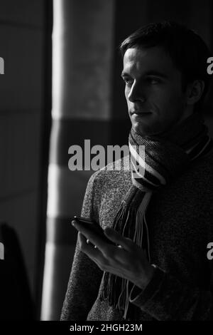 Ein junger, nachdenklicher, stilvoller Mann in einem modischen Pullover mit Schal steht neben dem Fenster und hält ein Telefon in den Händen und blickt auf den Bildschirm Stockfoto