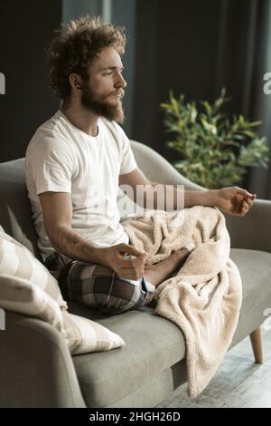 Junger entspannter Mann, der seinen täglichen Yoga auf dem Sofa in seiner Wohnung macht. Ein Mann mit betenden Händen und gefalteten Beinen, der in Lotus-Pose sitzt. Nahaufnahme. Hochwertige Fotos Stockfoto