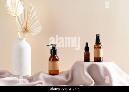 Set von Naturkosmetik in Bernstein-Glas-Verpackungen auf Satinstoff runden Podium und Vase mit getrockneten Blumen auf beigem Hintergrund. Stockfoto