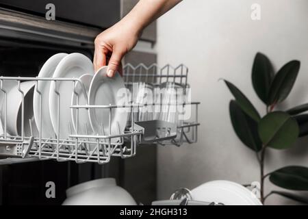 Nahaufnahme des weiblichen Handladens, das auf eine offene, automatische, integrierte Geschirrspülmaschine mit sauberen Utensilien in der modernen Wohnküche aufgetischt, ausgespült oder entladen wurde. Haushaltsleben