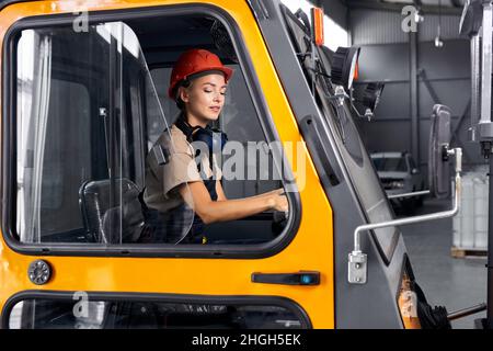 Arbeiterin fährt Lader im industriellen Containerlager, sieht selbstbewusst aus, gekleidet in Ingenieuruniform und orangefarbenem Helm, konzentriert Stockfoto