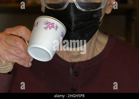 Ein Mann mit Schutzmaske und Schutzbrille mit einer Tasse Kaffee. Covid-19-Konzept. Stockfoto