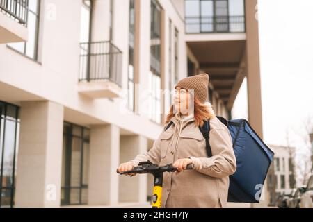 Low-Angle-Ansicht der weiblichen Kurierin mit großen thermischen Rucksack Reiten auf Elektroroller in der Stadt Straße wegschauen. Stockfoto