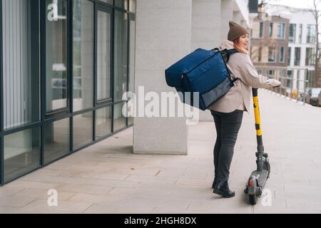 Low-Winkel-Rückansicht der weiblichen Kurier Lebensmittel Lieferung mit großen thermischen Rucksack zu Fuß mit Elektroroller in der Stadt Straße. Stockfoto