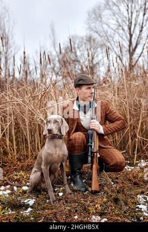 Aufmerksamer Rüde mit Weimaraner Hund sitzt in Büschen und Jagd nach einem Tier, in der wilden Natur. hunter und sein Hund auf der Suche nach Kleinwild. kaukasisch Stockfoto