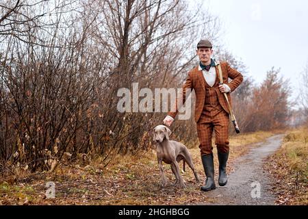 Selbstbewusster Mann Jäger mit Jagdhund Weimaraner Freund Porträt in ländlicher Umgebung während der Jagdsaison, schöner kaukasischer Kerl in braunem Anzug, zu Fuß Stockfoto