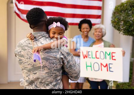 Lächelndes afroamerikanisches Mädchen, das den Vater eines Soldaten bei seiner Rückkehr umarmt, mit Familie im Hintergrund Stockfoto