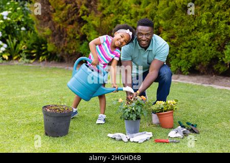 Porträt eines lächelnden afroamerikanischen Mädchens, das Pflanzen vom Vater gießt, der im Garten kniet Stockfoto