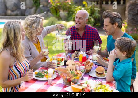 Fröhliche kaukasische Familie aus drei Generationen, die am Tisch im Garten zu Mittag essen kann Stockfoto