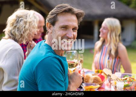 Porträt eines lächelnden kaukasischen Mannes, der zusammen mit der Familie am Tisch im Garten sitzt Stockfoto