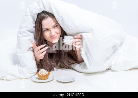 Guten Morgen. Weißes Schlafzimmer. Eine Frau liegt im Bett, hält eine Tasse heißes Getränk, ein Cupcake steht in der Nähe. Das Mädchen isst heimlich Frühstück. Stockfoto