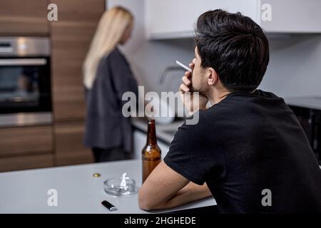 Paar mit Problemen mit Meinungsverschiedenheiten in der Küche, Mann sitzen Rauchen und Bier Alkohol trinken, während Frau kocht. Konzentrieren Sie sich auf Brunette Bartmann. Stockfoto