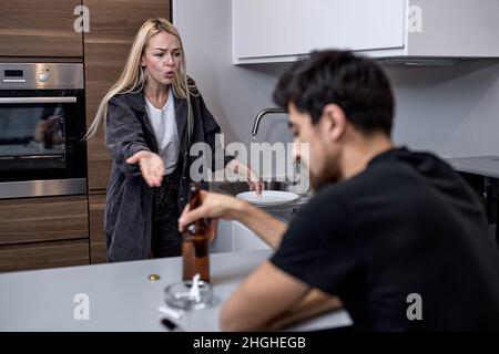 Paar mit Problemen mit Meinungsverschiedenheiten in der Küche, Mann sitzen Rauchen und Bier Alkohol trinken, während Frau kocht. Konzentriere dich auf Blonde Screaming Woman Stockfoto